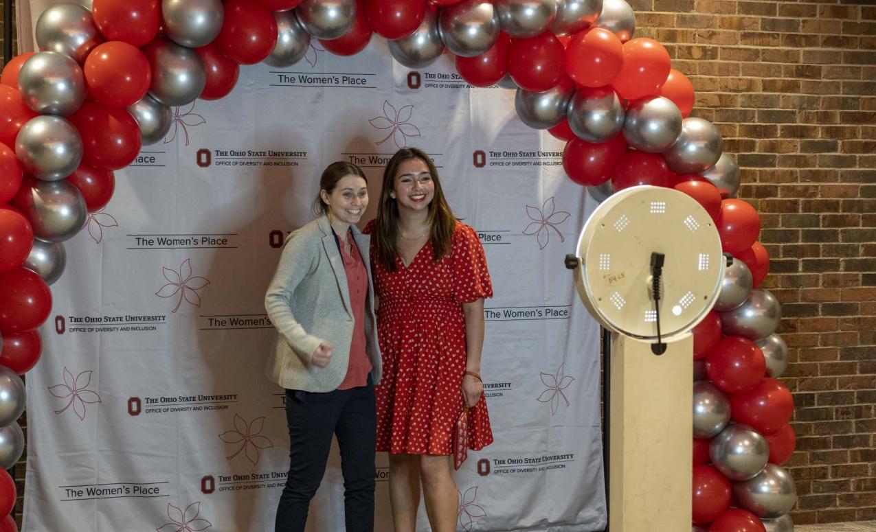Two young women standing, posing for photo under a scarlet and gray ballon arch.