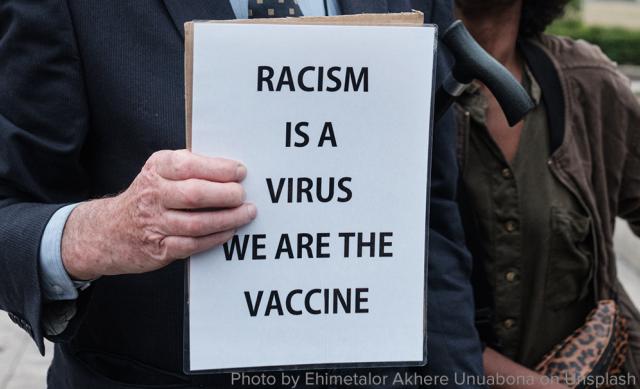 Hand holding sign stating "Racism is a virus, we are the vaccine"