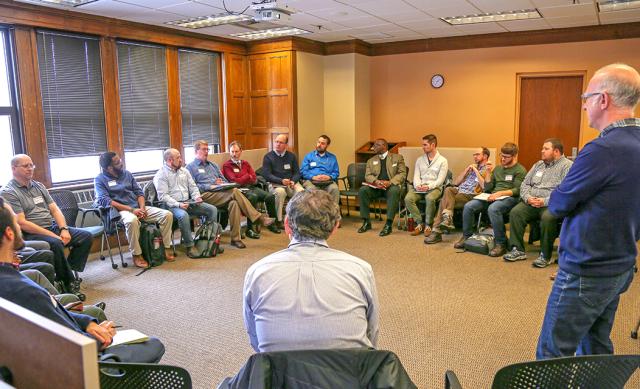 Circle of 20+ men in room listening to presentation by man standing