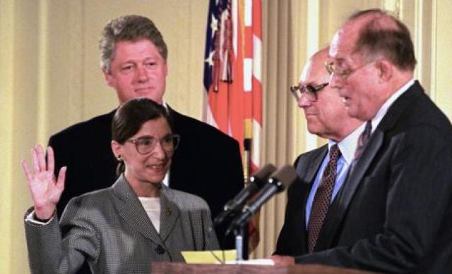 Ruth Bader Ginsberg holding up her right hand, with Bill Clinton behind her and two men in front of her, being sworn in as Supreme Court Justice.