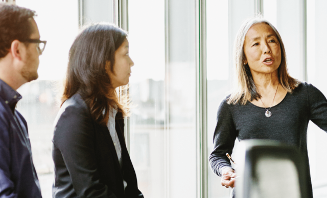Woman standing wearing business attire pointing speaking and gesturing while a man and woman listens