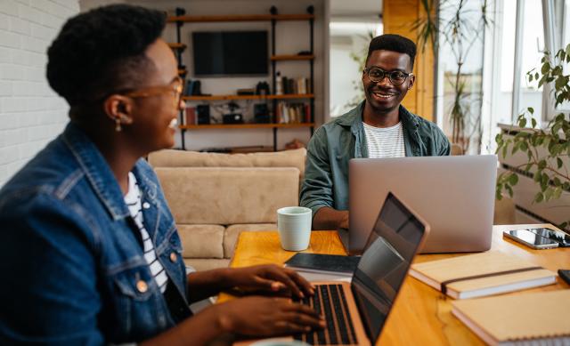 Black woman and man working from home, sitting at table together, each with laptop in front of them