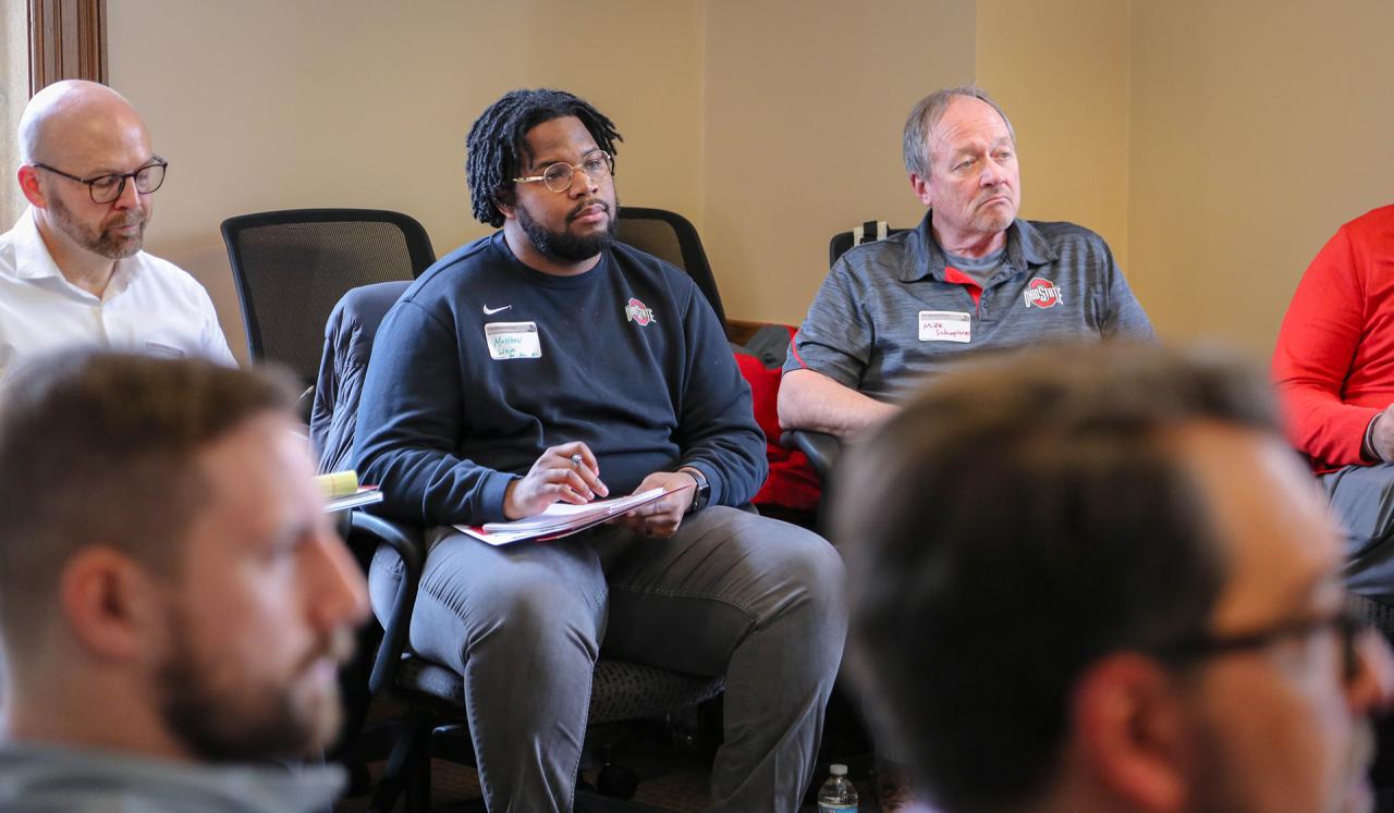 Three men wearing business casual clothing sitting next to each other listening intently with two men blurred in the foreground.