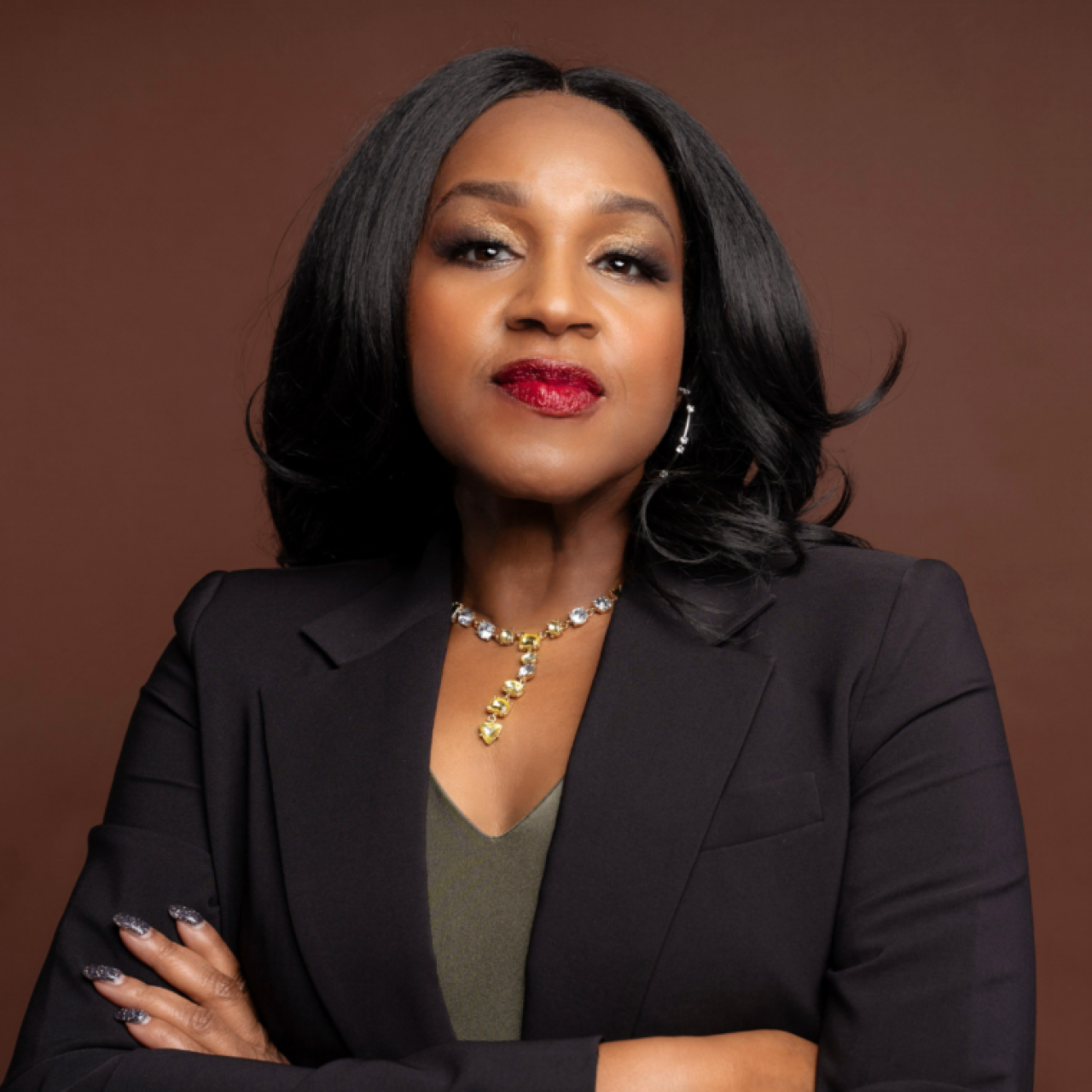 Elaine Richardson wearing a black blazer and red lipstick standing in front of a brown background