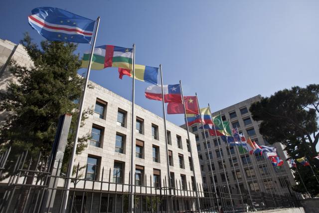 Multistory white building with many windows and a row of flags from different countries mounted on poles in front of the building