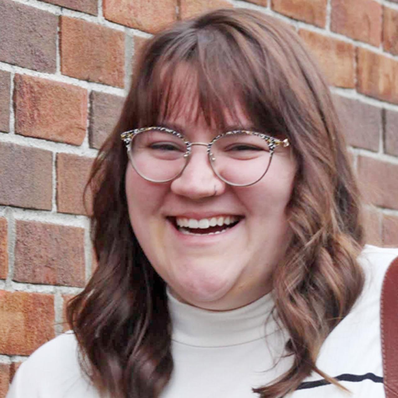 Young woman in white and black striped sweater with straight brown hair smiling and standing next to a brick wall