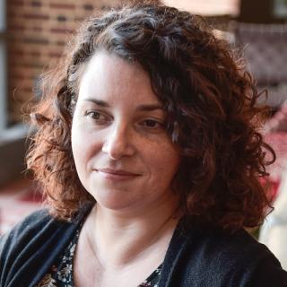 Portrait of Sara Childers wearing a black sweater and black and white flowered dress sitting in front of a brick wall