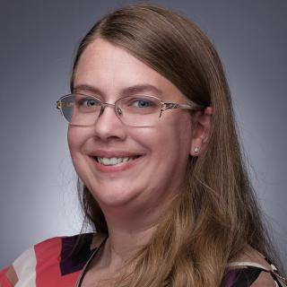 Photo of Angela Filous smiling standing in front of a gray background. 