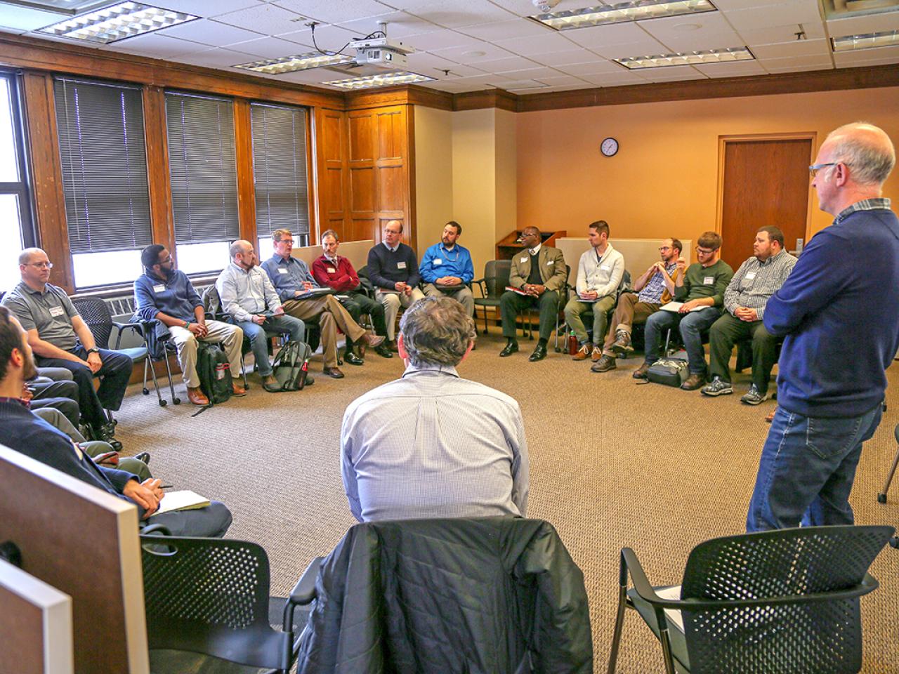 Circle of 20+ men in room listening to presentation by man standing