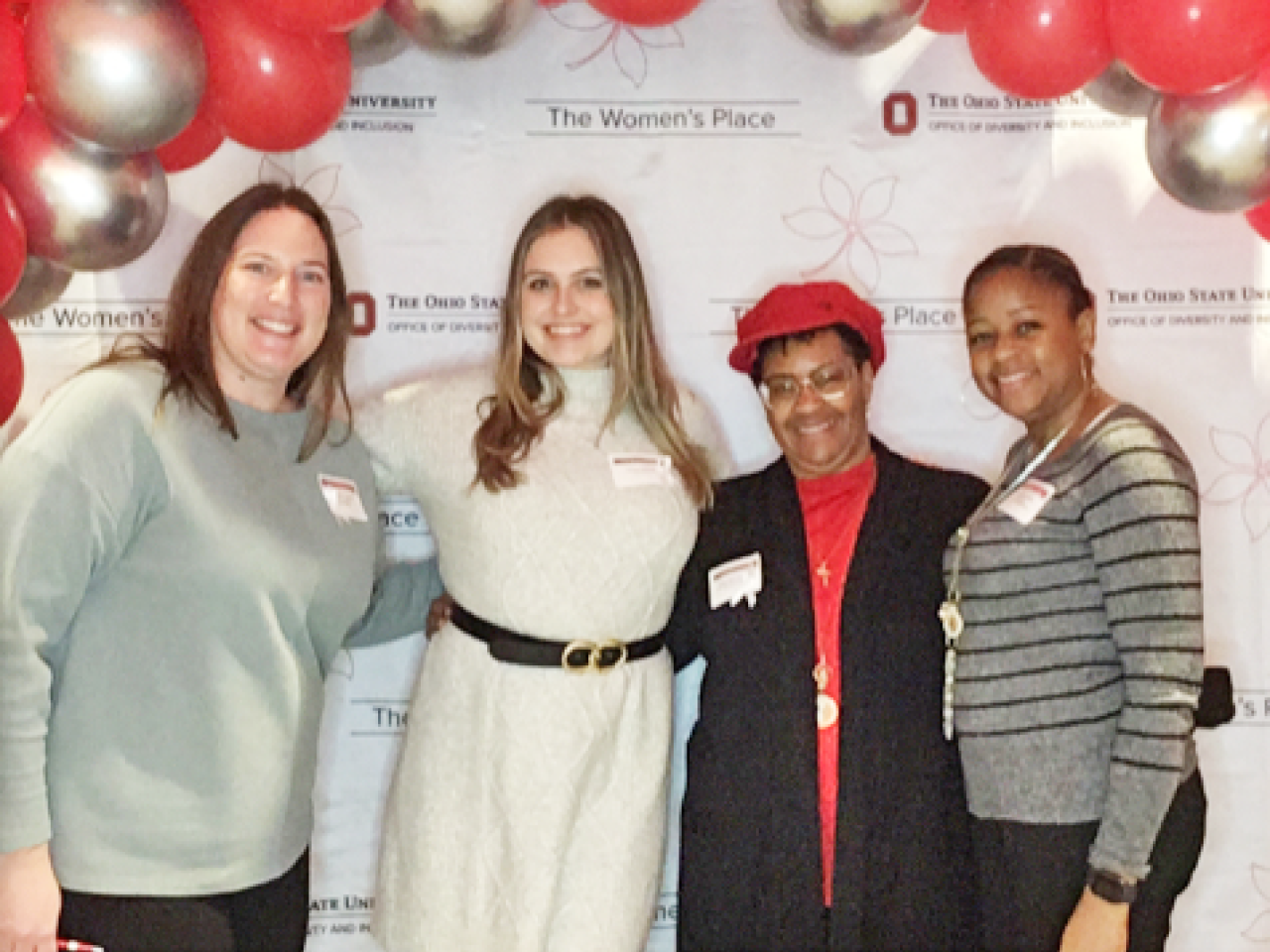 Four women in business casual dress stand under a scarlet and gray balloon arch and pose for the picture.