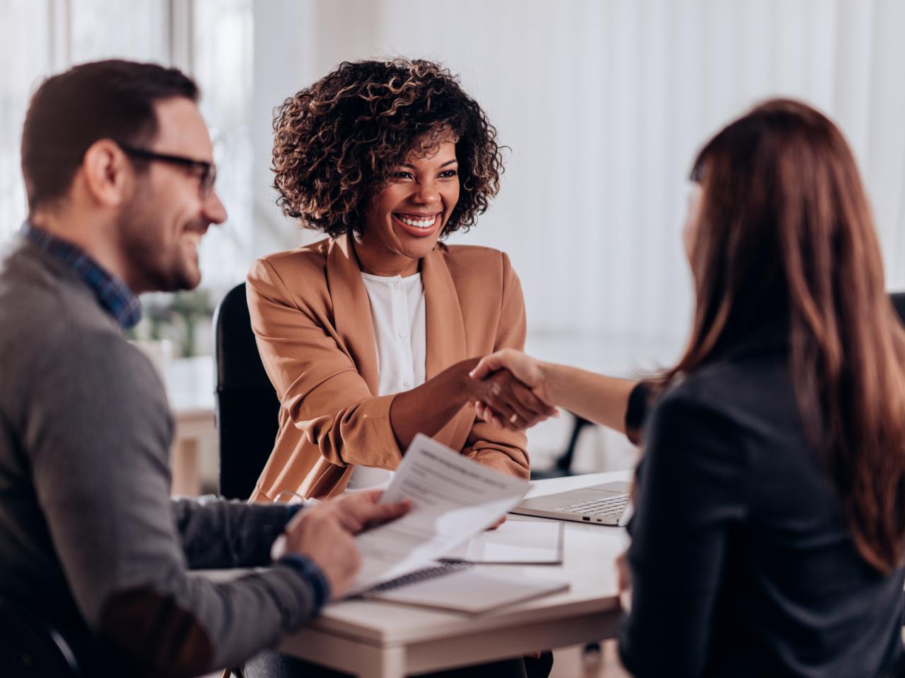 Group of three diverse people, two of which are shaking hands, in business clothing sitting together in a work setting