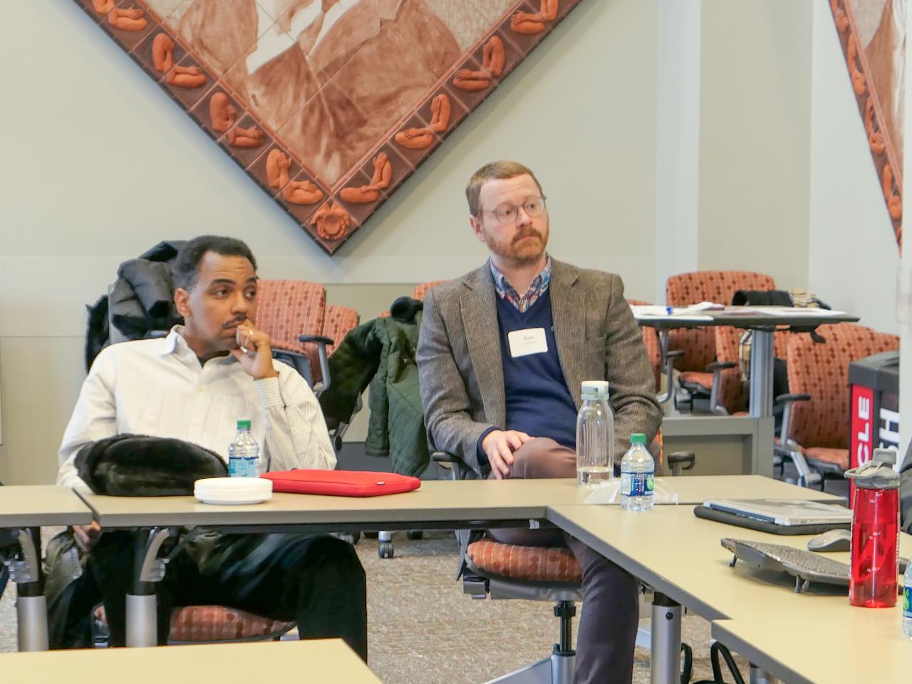 Three men sitting at a table, two wearing suits, one wearing button down shirt, listening to a lecture