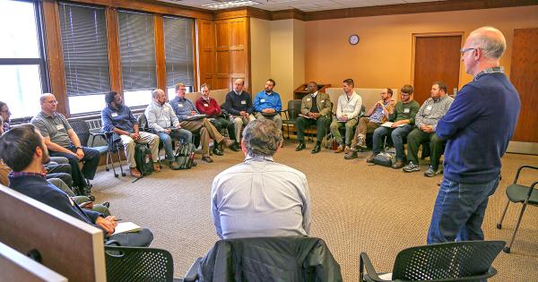 Circle of 20+ men in room listening to presentation by man standing