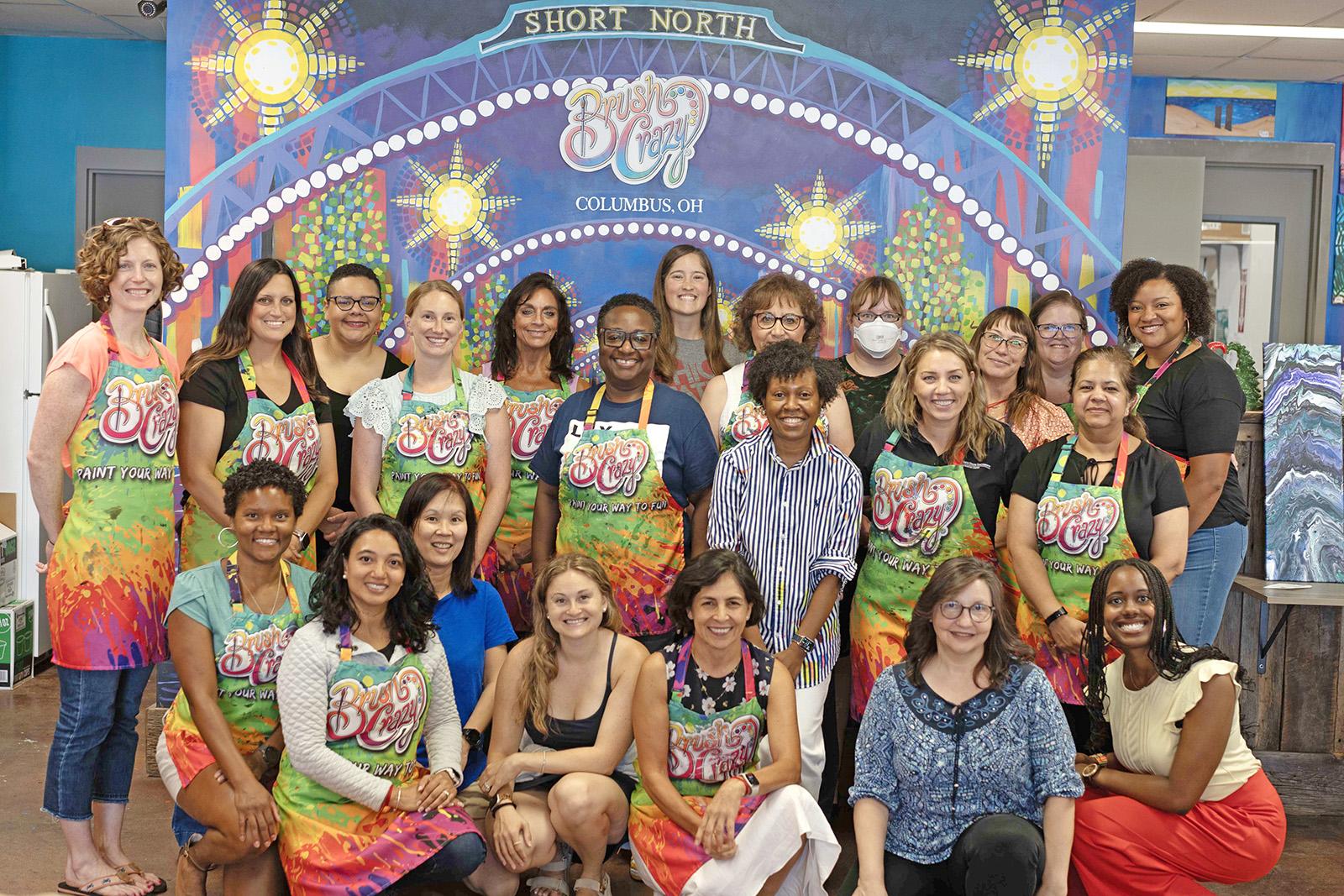 A diverse group of women wearing aprons that say "Brush Crazy" smiling and standing in front of a painting of the Short North in Columbus, Ohio. 