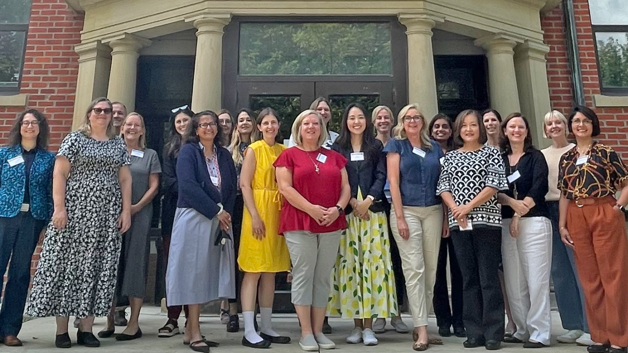 Diverse group of 20+ faculty women and one man wearing business casual clothes standing in front of building entrance
