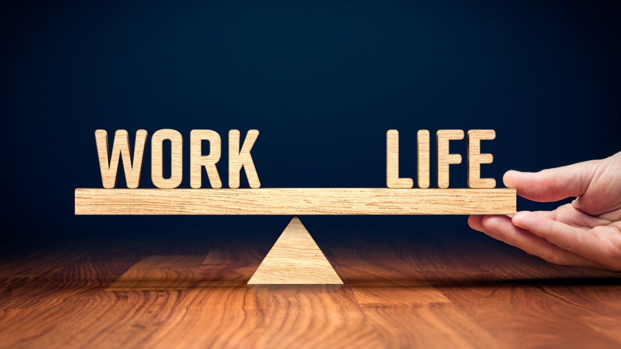 A wooden board balanced on a smaller wooden triangle. The board has letters on one end spelling out “work” and letters on the other end that spell out “life”. A hand is holding up the side that says “life”.  