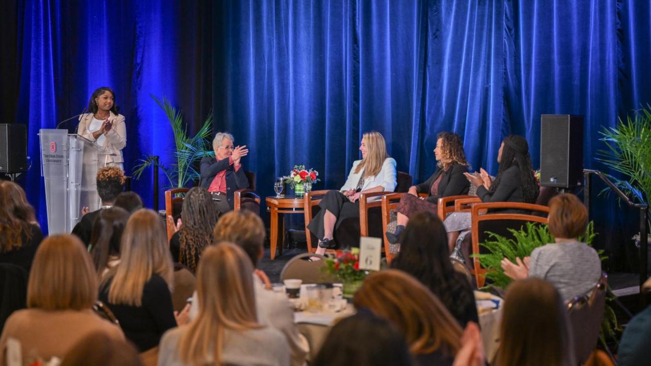 A speaker addresses an audience at the Women’s Leadership Symposium. Four panelists are seated on stage, listening.